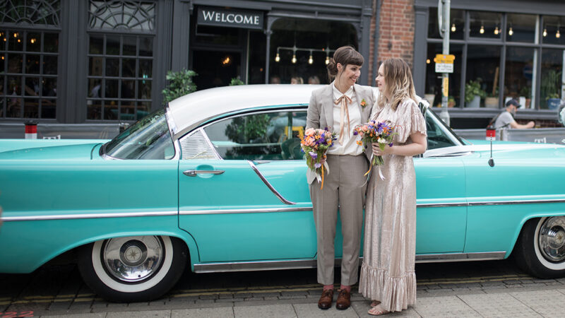 Two white women stand in front of the side door of a turquoise Cadillac. One is wearing a silver suit with a white shirt, the other a silver dress with ruffles across the front. They are both carrying flowers.