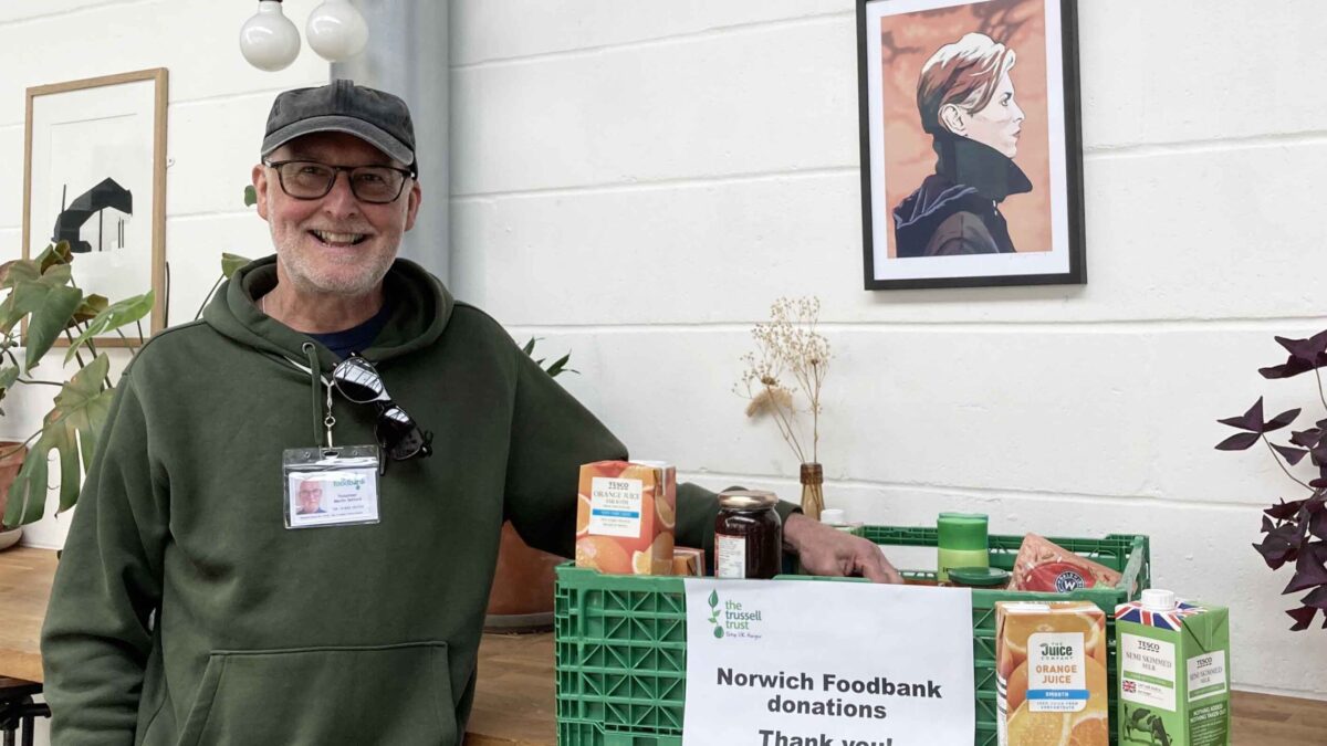 A smiling man wearing a green hoodie and cap stands next to a crate of donated food items with a sign on the crate that reads Norwich Foodbank donations. Thank you!