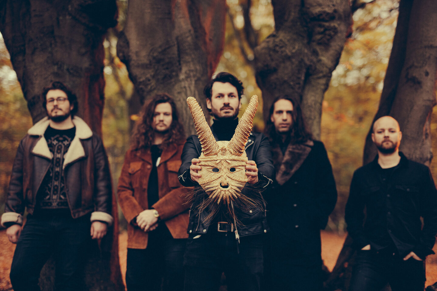 An out of focus group of 5 men stand in a row against a woodland background. The middle man holds up a white rabbit mask. An out of focus group of 5 men stand in a row against a woodland background. The middle man holds up a white rabbit mask.