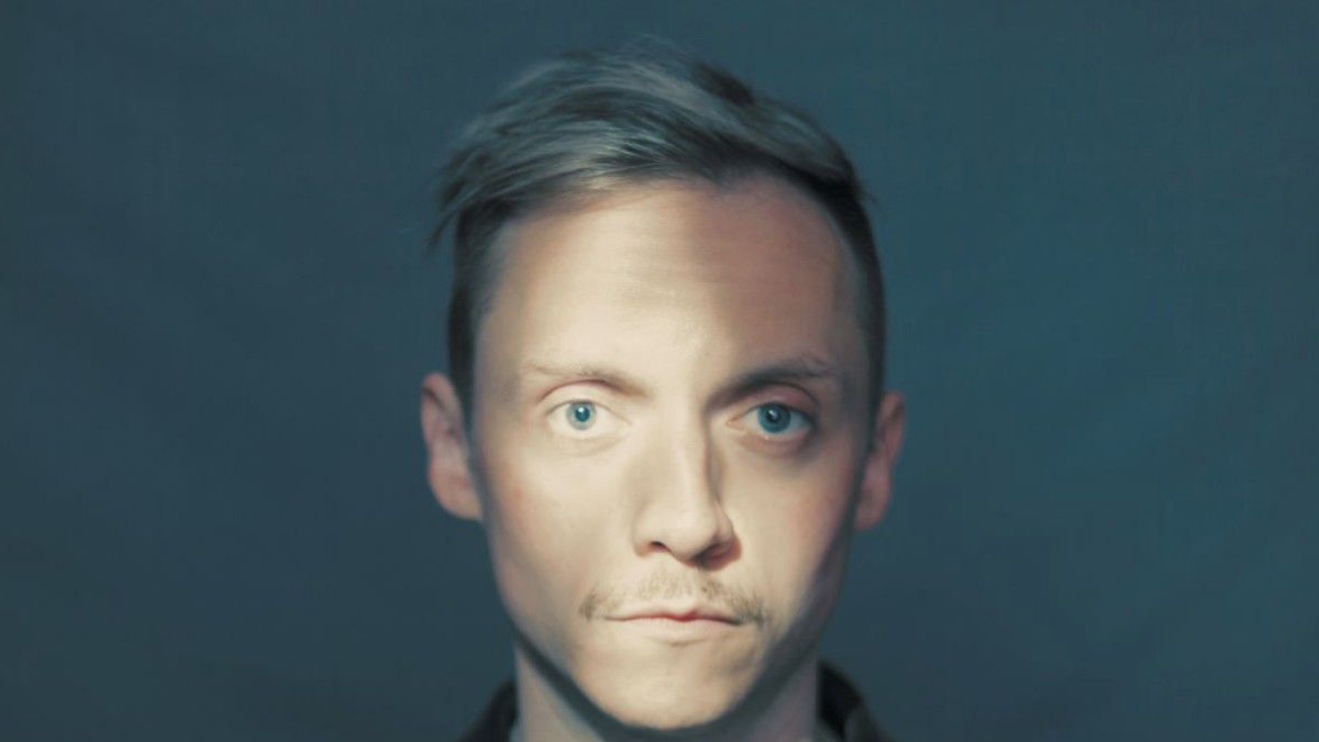 A close up of a man with short blond / grey hair staring directly at the camera against a blue / grey background.