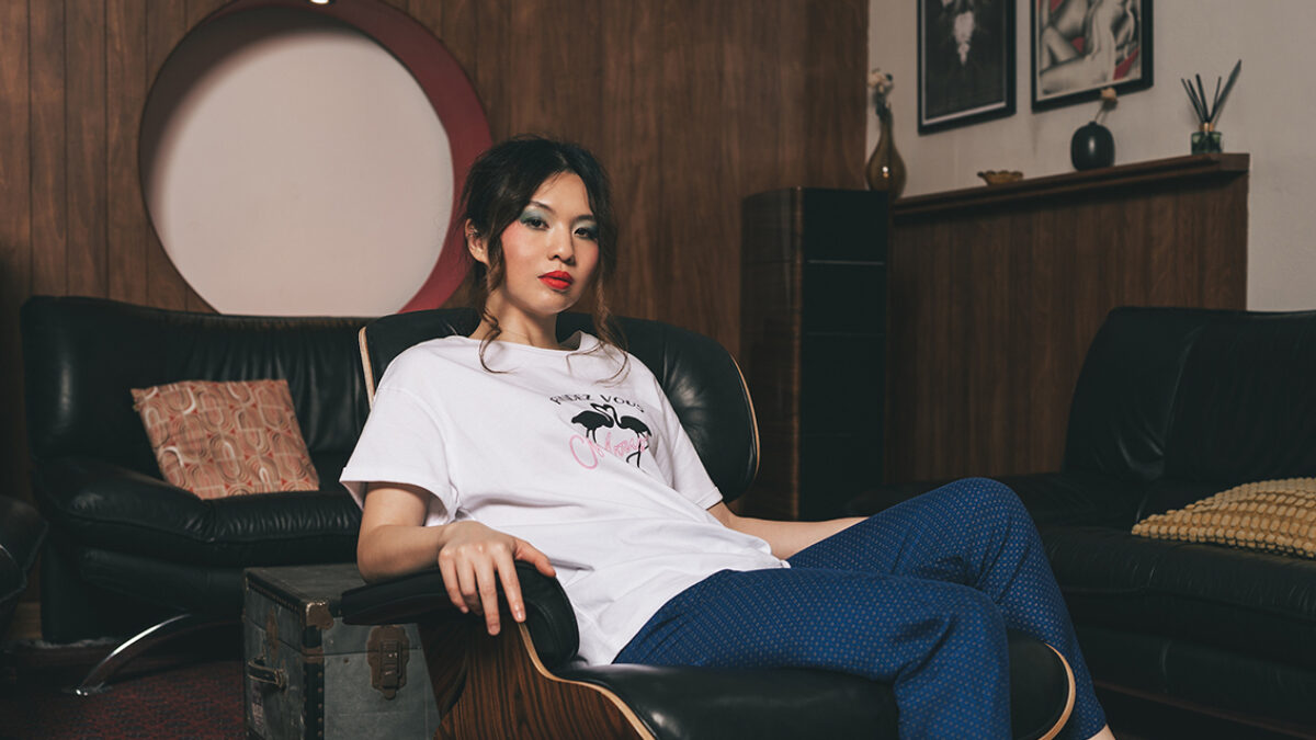 A women wearing a white t-shirt and blue trousers is sat on a black leather chair, in front of a sofa and wooden wall. She has dark hair strands falling to each side of her face.