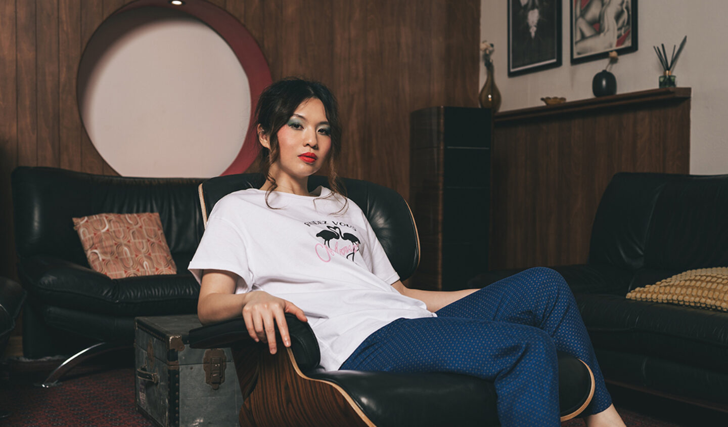 A women wearing a white t-shirt and blue trousers is sat on a black leather chair, in front of a sofa and wooden wall. She has dark hair strands falling to each side of her face.