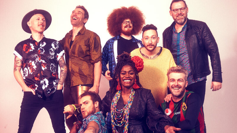 The 8 members of the London Afrobeat Collective are gathered together in form to a plain background.