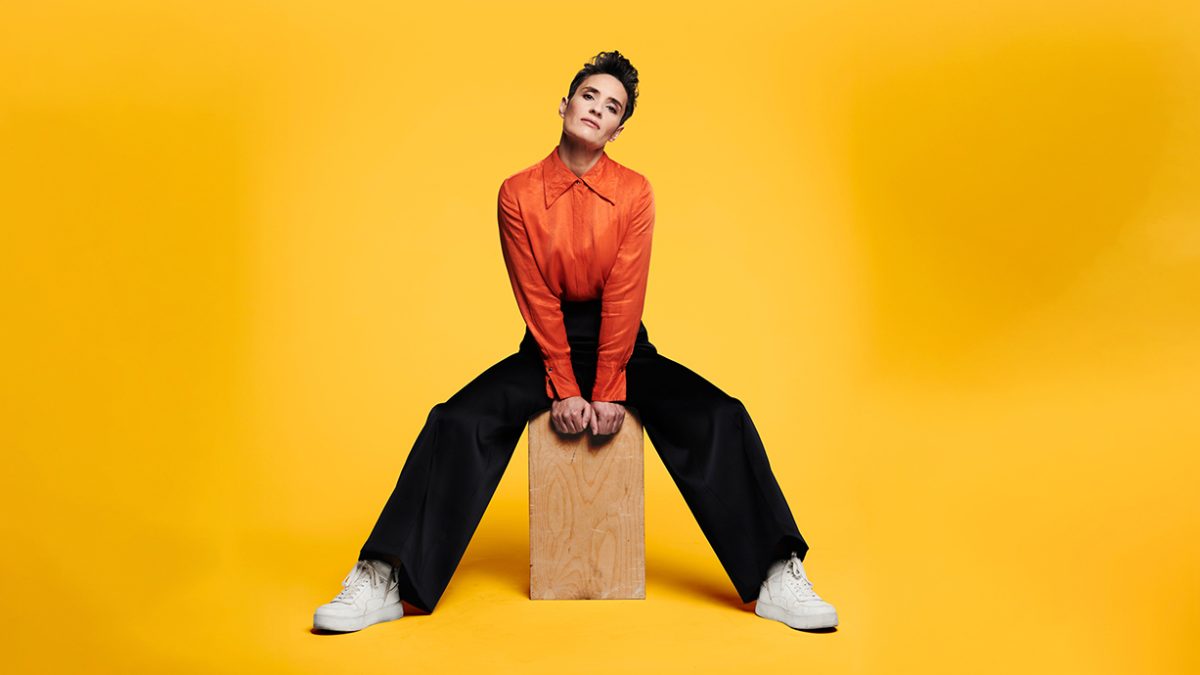A person, Jen Brister, sits on a box on a yellow background. They are in black trousers and an orange shirts, they have short dark hair and are looking forwards.