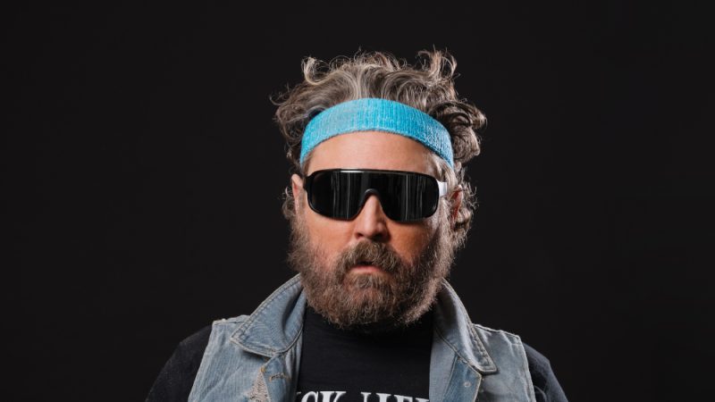 A head and shoulders of a person on the black background. They have short brown hair, a beard and wearing sunglasses, blue headband, and a denim vest. A head and shoulders of a person on the black background. They have short brown hair, a beard and wearing sunglasses, blue headband, and a denim vest.