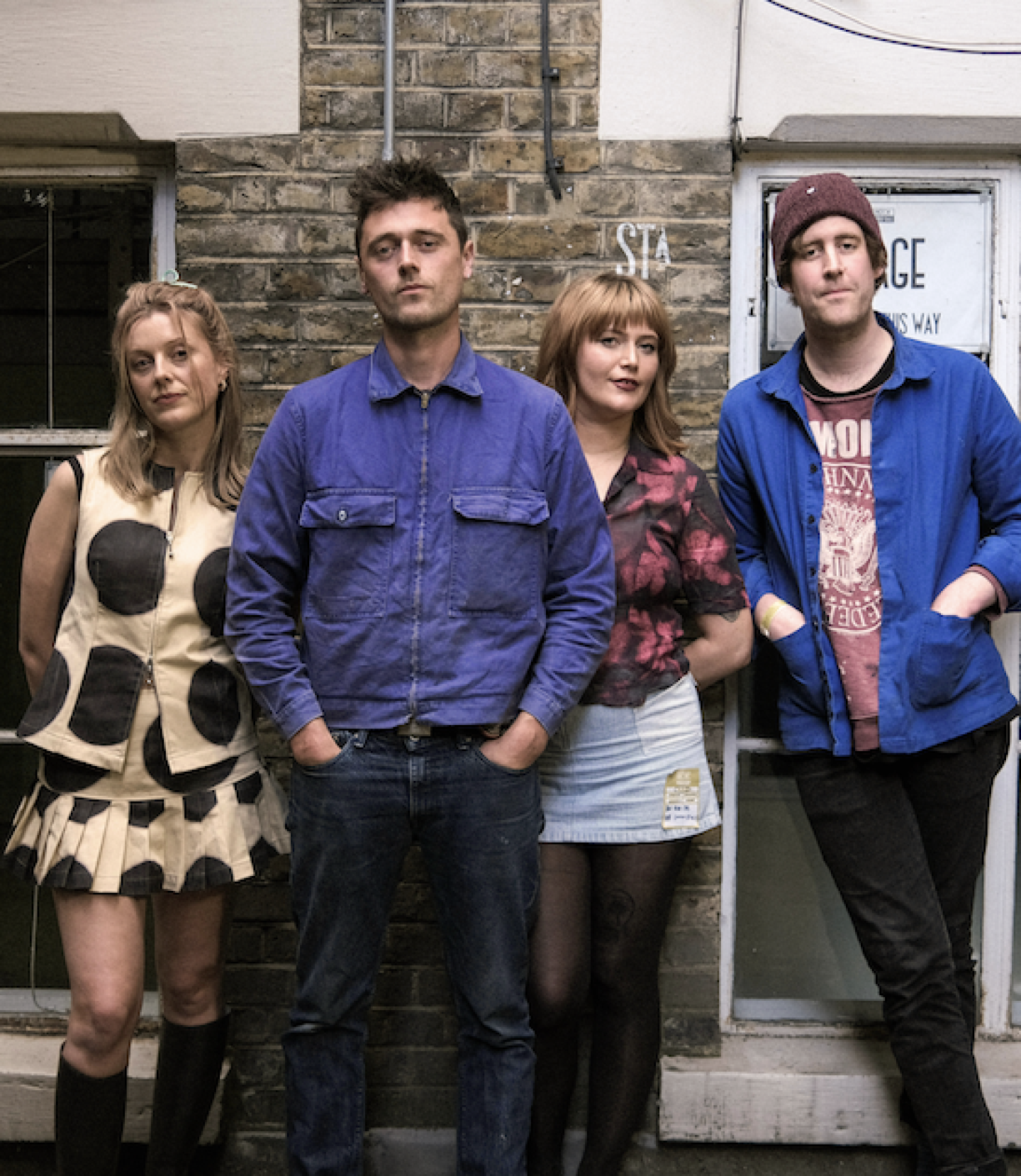Four people standing against a brick wall, two women and two men, dressed in casual and retro-style clothing. Four people standing against a brick wall, two women and two men, dressed in casual and retro-style clothing.