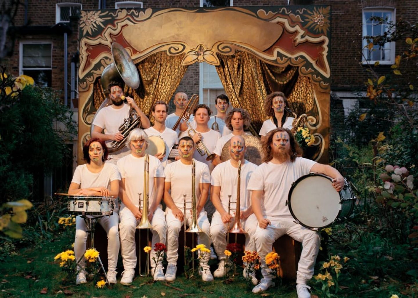 A group of 12 people arranged in front of a theatre backdrop in the garden. They are all wearing white, holding their brass and drum instruments and have a handprint face paint on their face. A group of 12 people arranged in front of a theatre backdrop in the garden. They are all wearing white, holding their brass and drum instruments and have a handprint face paint on their face.