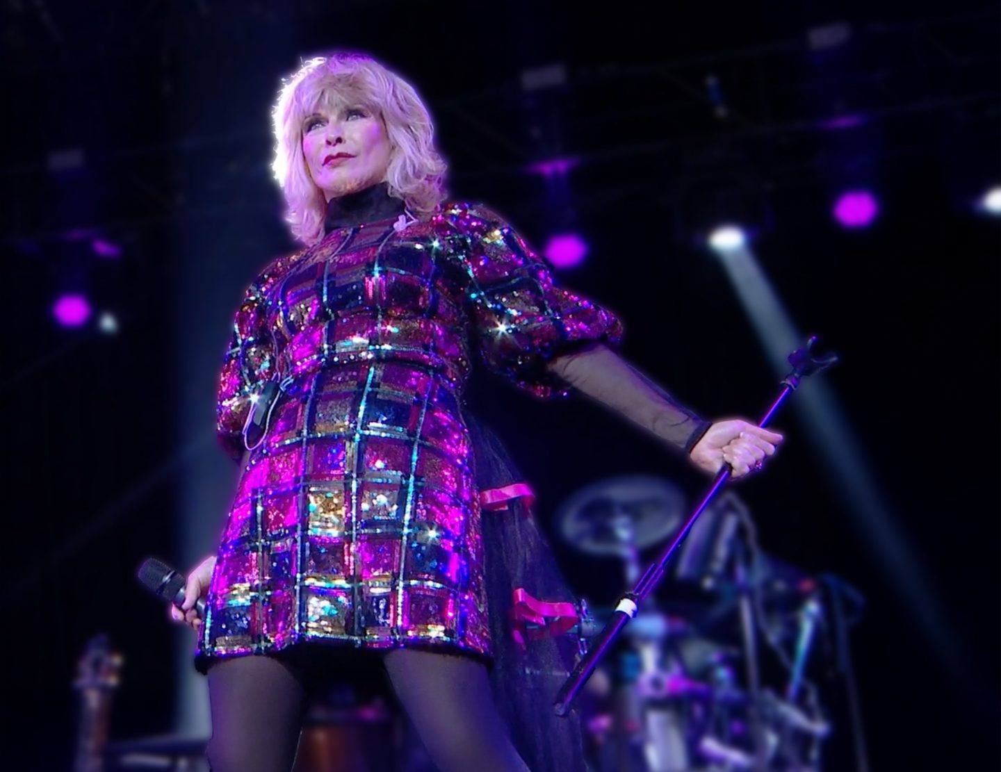 Toyah stands on stage with purple and white lights behind her. She has blonde hair to her shoulders and is wearing a shiny dress. Toyah stands on stage with purple and white lights behind her. She has blonde hair to her shoulders and is wearing a shiny dress.