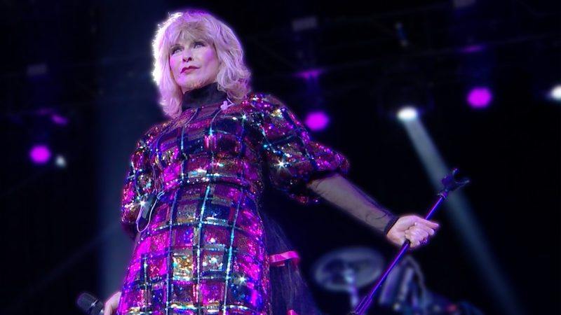 Toyah stands on stage with purple and white lights behind her. She has blonde hair to her shoulders and is wearing a shiny dress. Toyah stands on stage with purple and white lights behind her. She has blonde hair to her shoulders and is wearing a shiny dress.