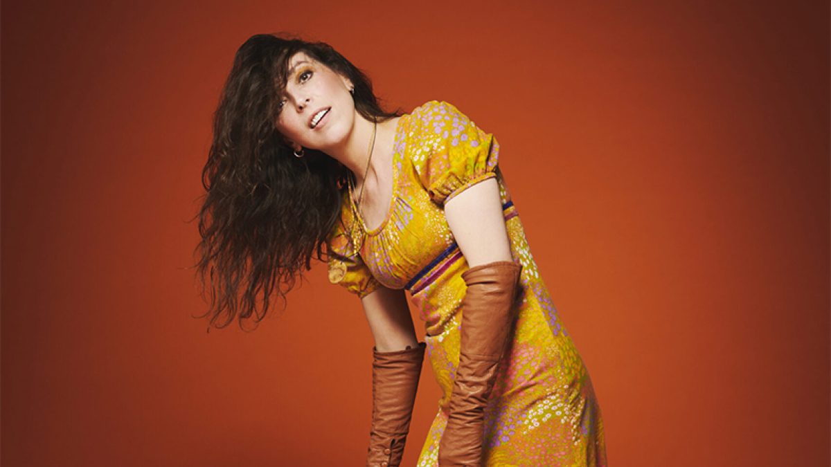 A figure on a dark orange background. They have long dark hair which is hanging to one side of their face, they are wearing a long flowery yellow dress and orange gloves to the elbow.