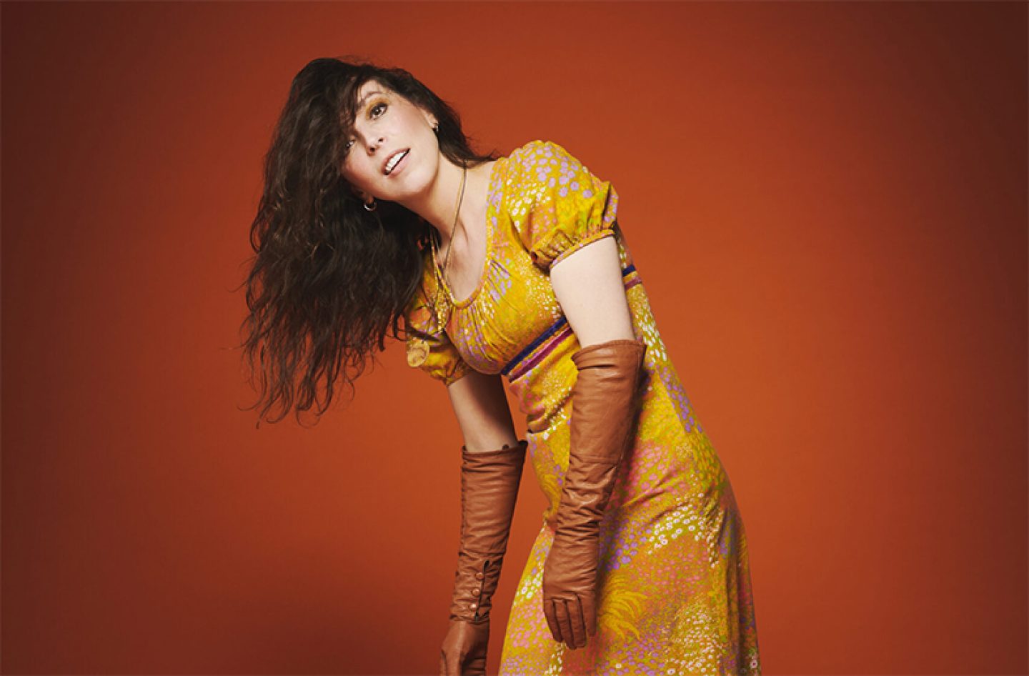 A figure on a dark orange background. They have long dark hair which is hanging to one side of their face, they are wearing a long flowery yellow dress and orange gloves to the elbow. A figure on a dark orange background. They have long dark hair which is hanging to one side of their face, they are wearing a long flowery yellow dress and orange gloves to the elbow.