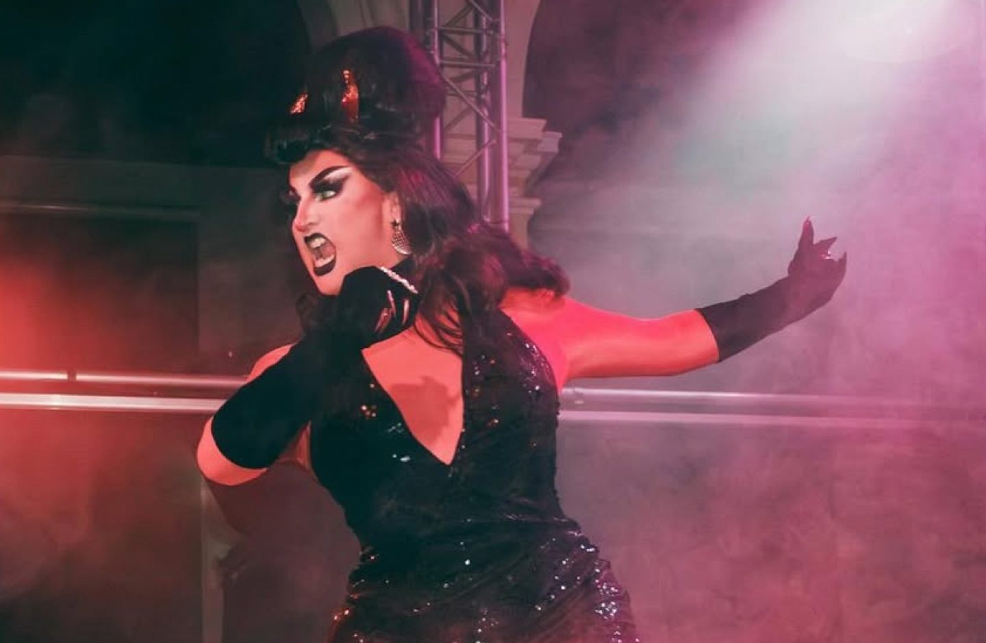 A head and shoulders image of a drag performer dressed in a V neck black top and long black gloves, with their hair is in a beehive style. They are looking to one side with their mouth open, their arms are held to one side as though they are in the middle of a movement. A head and shoulders image of a drag performer dressed in a V neck black top and long black gloves, with their hair is in a beehive style. They are looking to one side with their mouth open, their arms are held to one side as though they are in the middle of a movement.