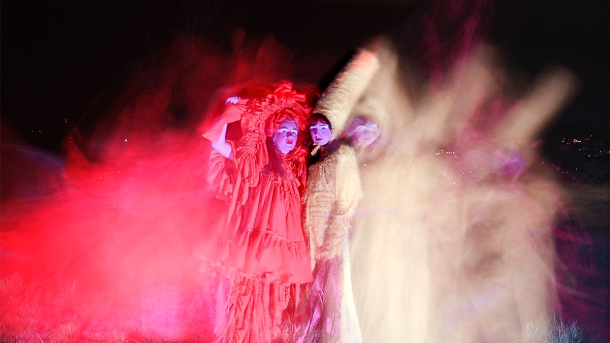 Two figures standing on grass at night. Wearing a red dress and head piece made of rippled fabric, the other a tall yellow hat and matching long yellow dress. There is a blur of colour around them, created by a low shutter speed on the camera.