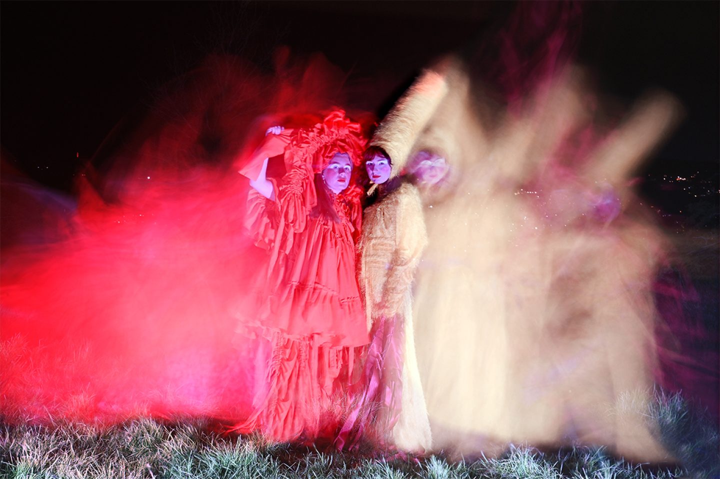 Two figures standing on grass at night. Wearing a red dress and head piece made of rippled fabric, the other a tall yellow hat and matching long yellow dress. There is a blur of colour around them, created by a low shutter speed on the camera. Two figures standing on grass at night. Wearing a red dress and head piece made of rippled fabric, the other a tall yellow hat and matching long yellow dress. There is a blur of colour around them, created by a low shutter speed on the camera.