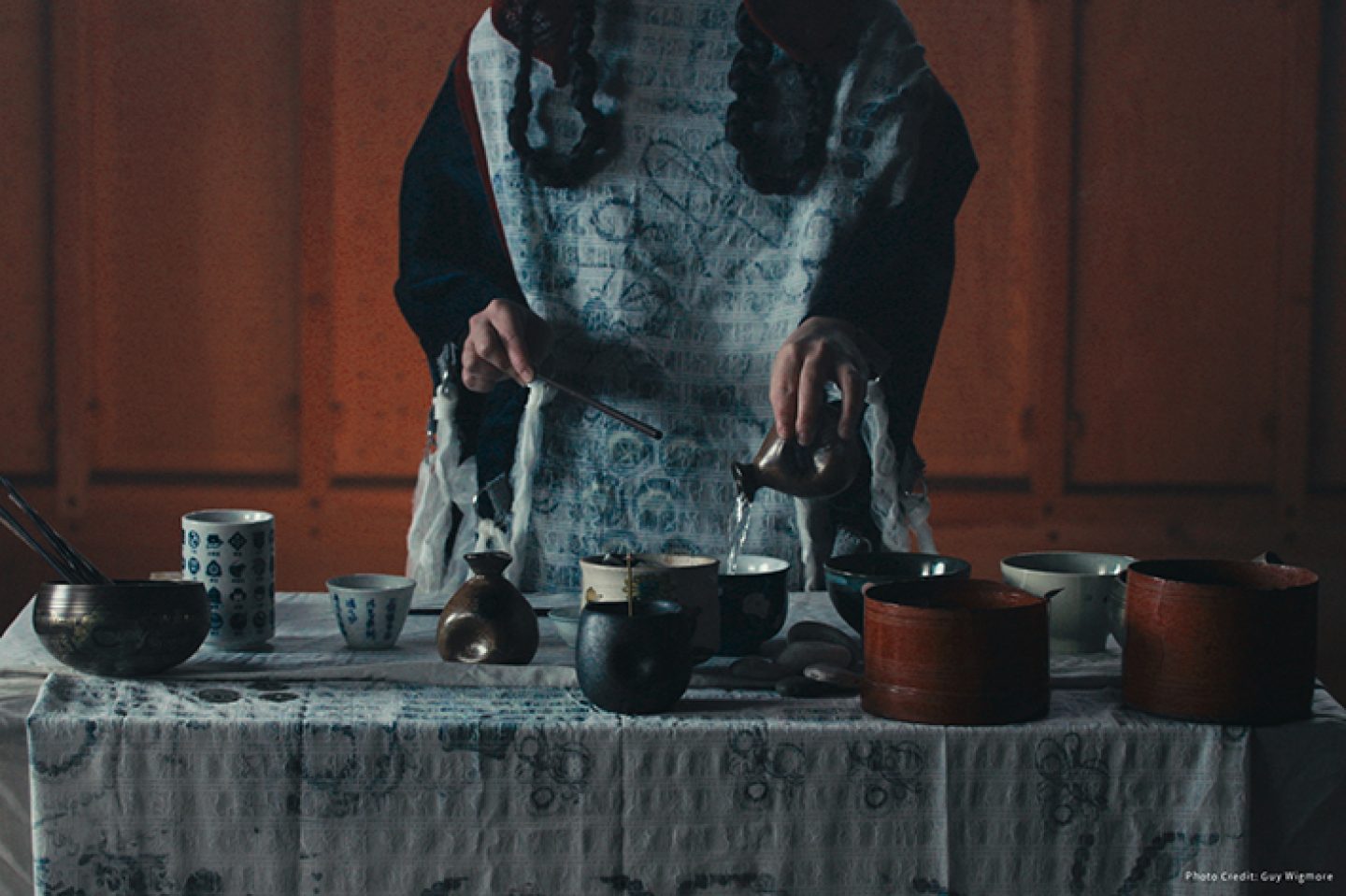 A table scene with many metal and ceramic receptacles on it. A torso of a figure is behind the table, one hand is pouring a liquid into one of the bowls. A table scene with many metal and ceramic receptacles on it. A torso of a figure is behind the table, one hand is pouring a liquid into one of the bowls.