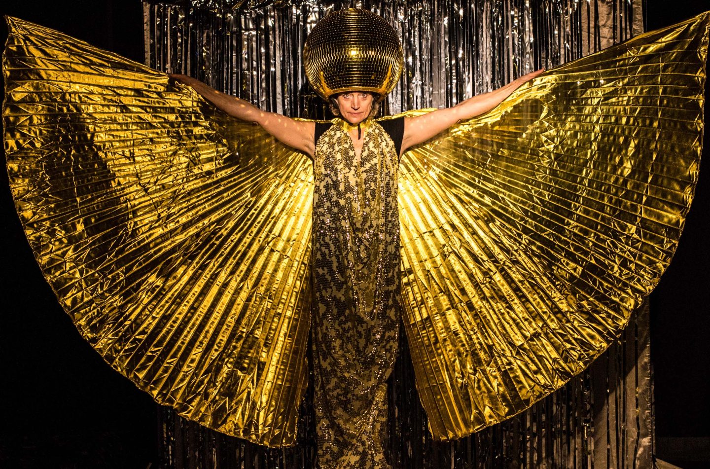 A figure stands in a gold dress wearing a gold disco ball on their head. Their arms are outstretched and raised creating wing shapes with gold material. Behind them is the cartoon of silver ribbon strips A figure stands in a gold dress wearing a gold disco ball on their head. Their arms are outstretched and raised creating wing shapes with gold material. Behind them is the cartoon of silver ribbon strips
