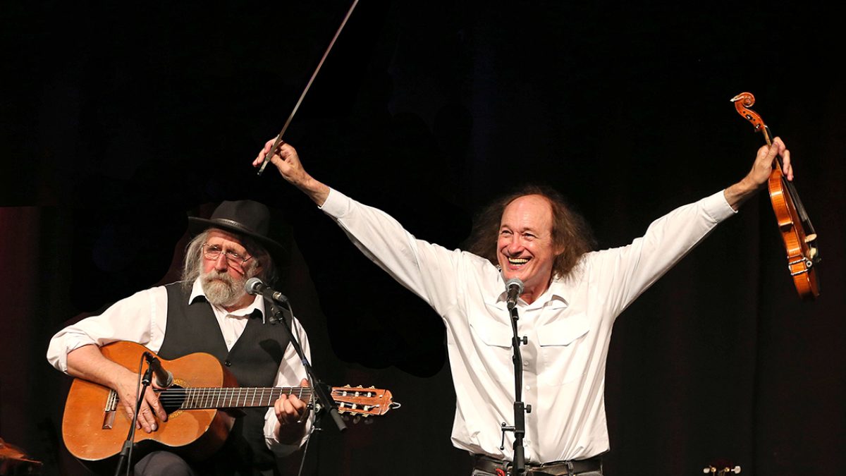 An image of two people in front of microphones. They are both wearing dark trousers and white shirts. One is holding a guitar and the other it's holding both arms in the air, they whipping a bow and violin.