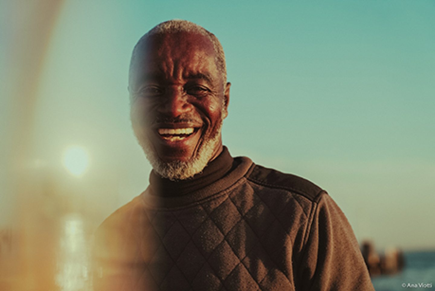A head and shoulders image of a person looking towards the camera smiling, with a low sun behind them. They have short grey hair and beard and they're wearing a dark high neck sweatshirt. A head and shoulders image of a person looking towards the camera smiling, with a low sun behind them. They have short grey hair and beard and they're wearing a dark high neck sweatshirt.