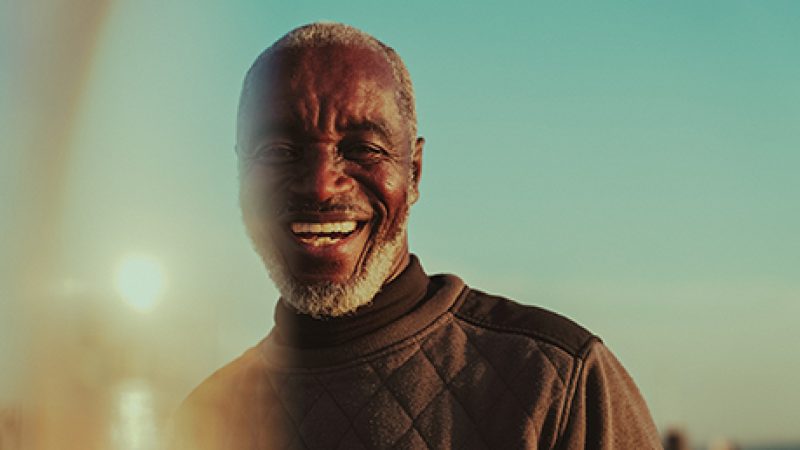 A head and shoulders image of a person looking towards the camera smiling, with a low sun behind them. They have short grey hair and beard and they're wearing a dark high neck sweatshirt. A head and shoulders image of a person looking towards the camera smiling, with a low sun behind them. They have short grey hair and beard and they're wearing a dark high neck sweatshirt.