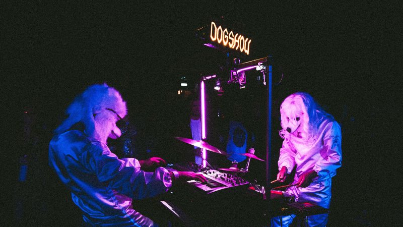 Image of two figures in shiny suits with white fluffy dogs heads. They are either side of a collection of instruments and lights. Above them is a sign that reads dogshow.