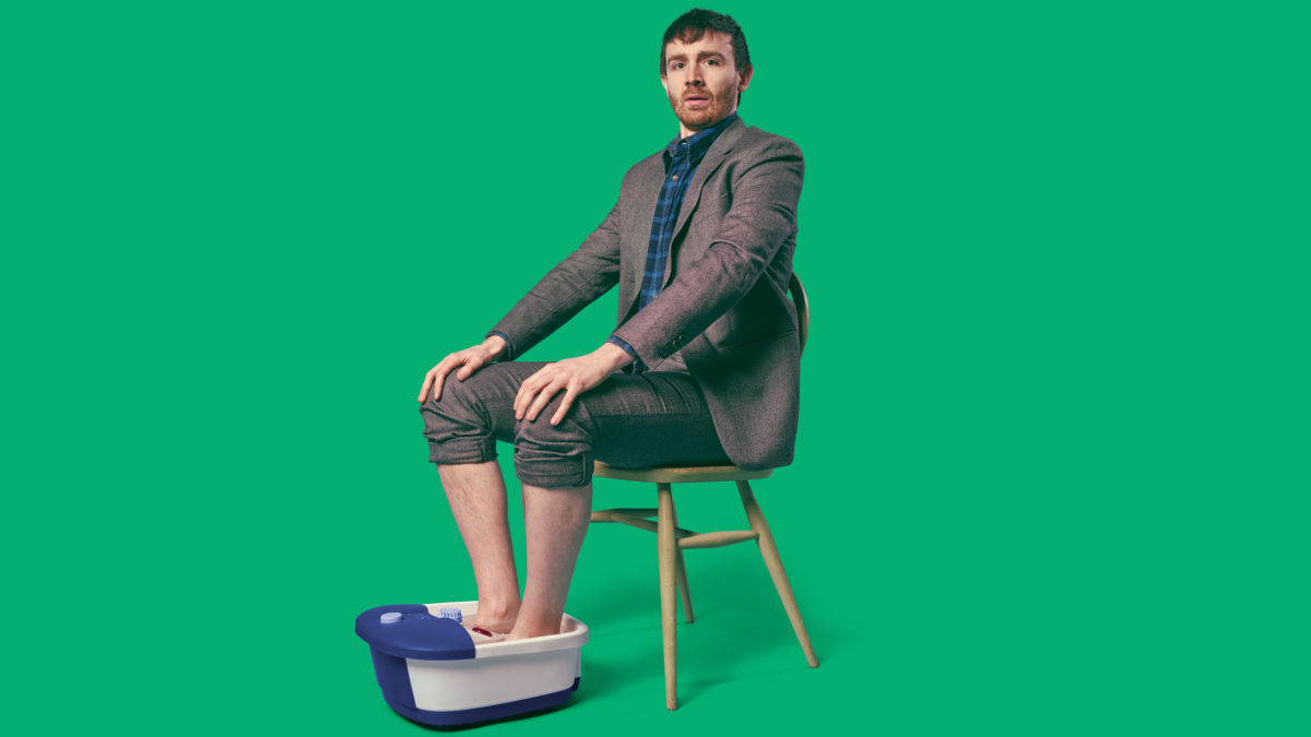 A person sits on a chair with their feet in a foot spa. They are wearing a suit and checked shirt and have short brown hair. The background is bright green.