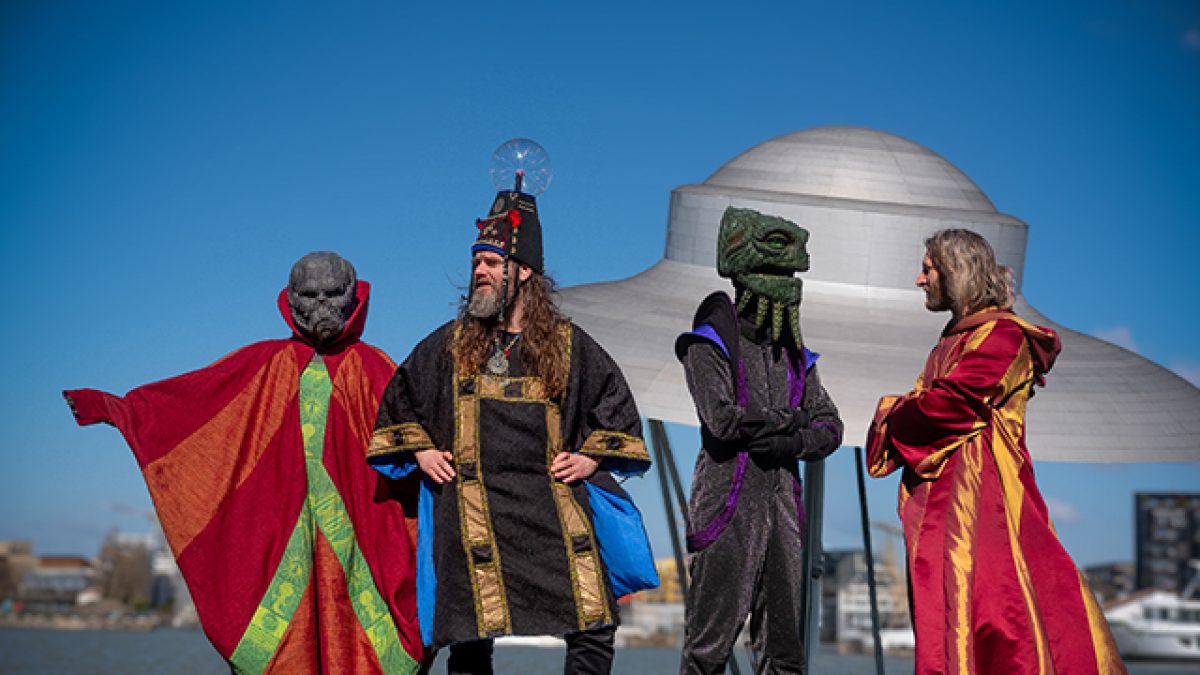 Four figures stood in front of a sculpture shaped like a UFO. Two of the figures have alien heads, one is wearing a large hat with a plasma ball on it.
