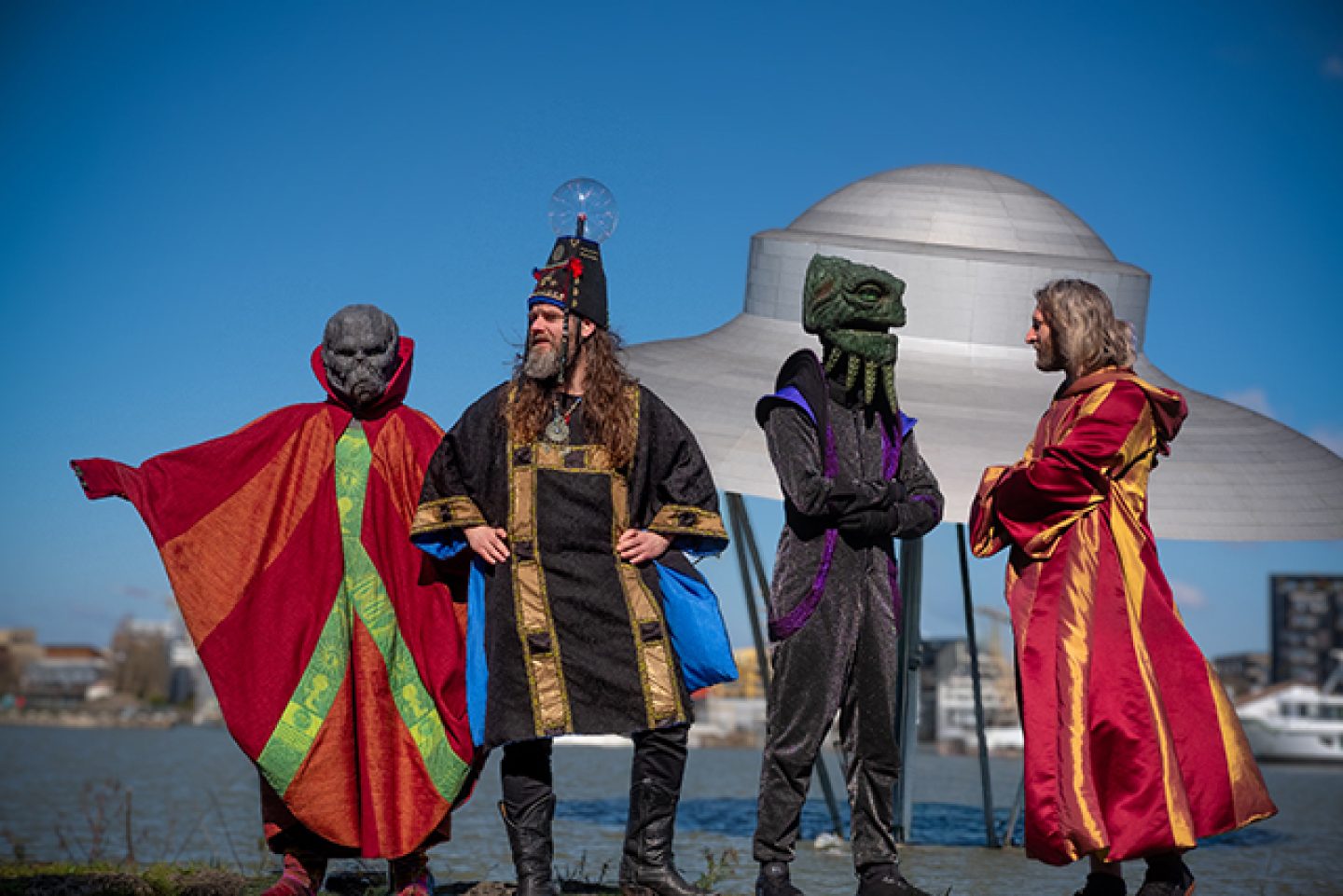 Four figures stood in front of a sculpture shaped like a UFO. Two of the figures have alien heads, one is wearing a large hat with a plasma ball on it. Four figures stood in front of a sculpture shaped like a UFO. Two of the figures have alien heads, one is wearing a large hat with a plasma ball on it.