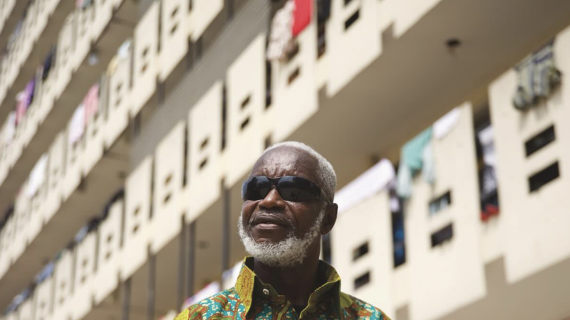 An older man with gray hair and a beard wearing sunglasses and a brightly patterned shirt stands in front of a tall apartment building with balconies and hanging laundry