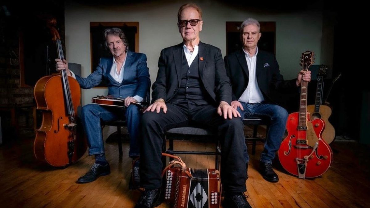 Three people sat on chairs wearing suits. Each has an instrument next to them, a double bass, and accordion, an electric guitar.