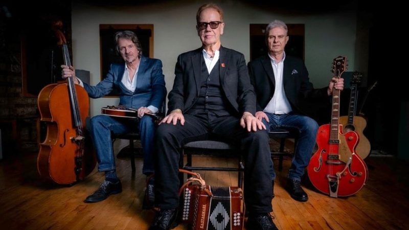 Three people sat on chairs wearing suits. Each has an instrument next to them, a double bass, and accordion, an electric guitar. Three people sat on chairs wearing suits. Each has an instrument next to them, a double bass, and accordion, an electric guitar.