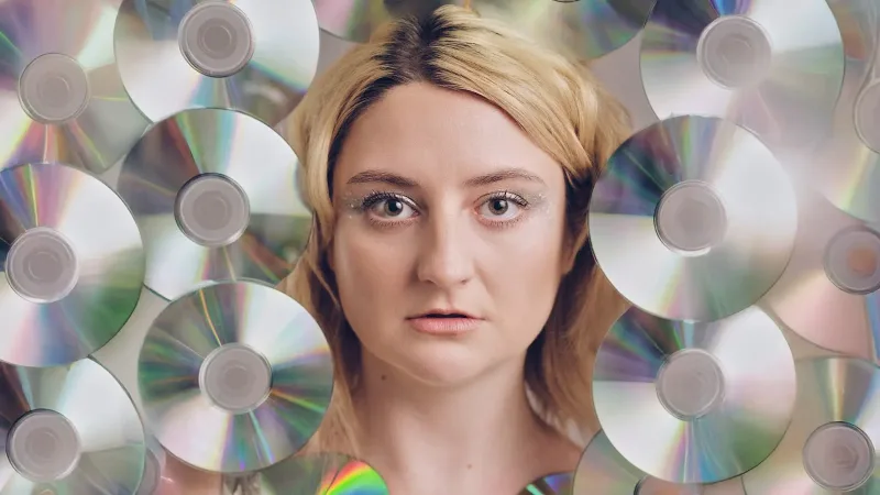 A woman with blonde hair and glittery eye makeup looks directly at the camera, surrounded by reflective CDs. A woman with blonde hair and glittery eye makeup looks directly at the camera, surrounded by reflective CDs.