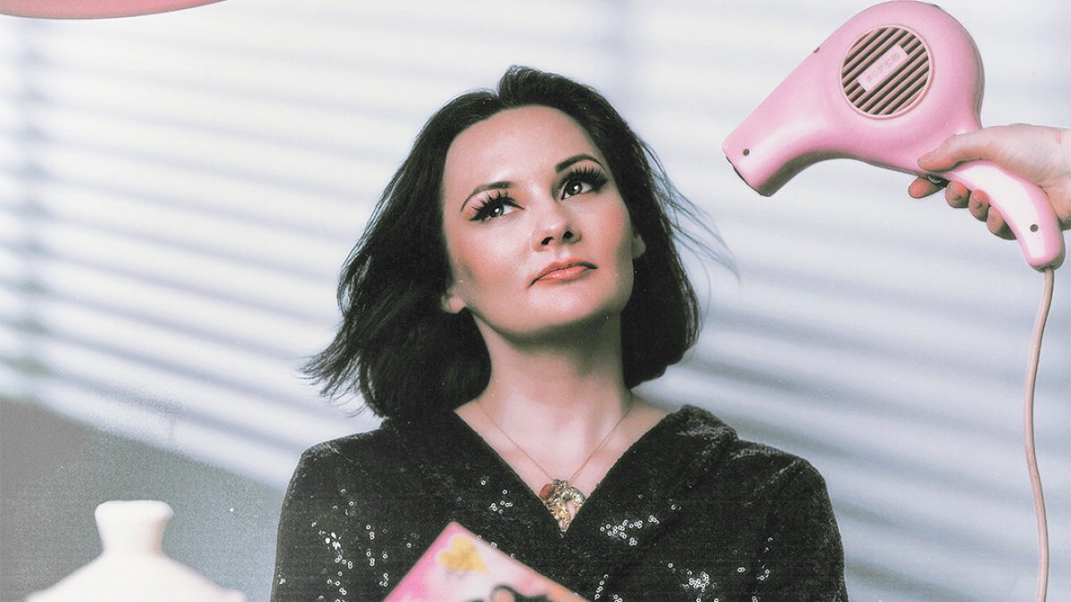 A woman with dark hair sits at a table holding a book, while someone off-camera points a pink hairdryer at her; teacups, a teapot, and other objects are arranged in the foreground.