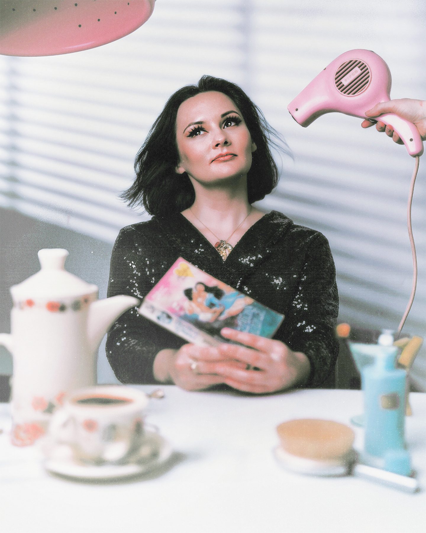 A woman with dark hair sits at a table holding a book, while someone off-camera points a pink hairdryer at her; teacups, a teapot, and other objects are arranged in the foreground. A woman with dark hair sits at a table holding a book, while someone off-camera points a pink hairdryer at her; teacups, a teapot, and other objects are arranged in the foreground.