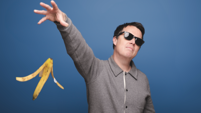 A man wearing sunglasses and a gray jacket playfully tosses a banana peel standing against a blue background. A man wearing sunglasses and a gray jacket playfully tosses a banana peel standing against a blue background.