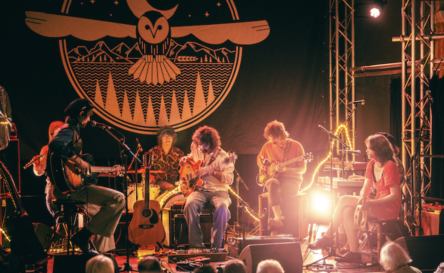 A band performs on stage with guitars, drums, and a tambourine in front of a large backdrop featuring an owl design A band performs on stage with guitars, drums, and a tambourine in front of a large backdrop featuring an owl design