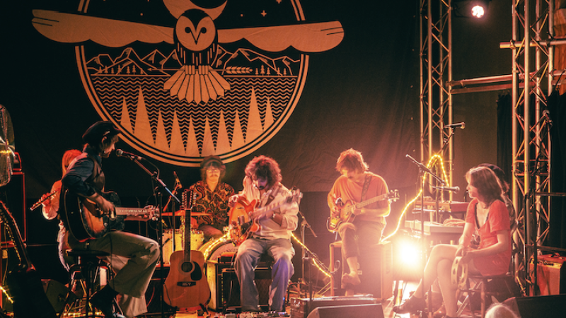 A band performs on stage with guitars, drums, and a tambourine in front of a large backdrop featuring an owl design A band performs on stage with guitars, drums, and a tambourine in front of a large backdrop featuring an owl design