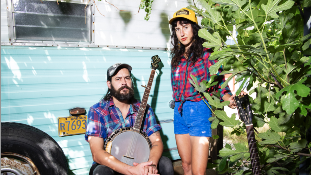 A man with a banjo and woman with a guitar pose by a vintage camper, wearing plaid shirts and trucker hats