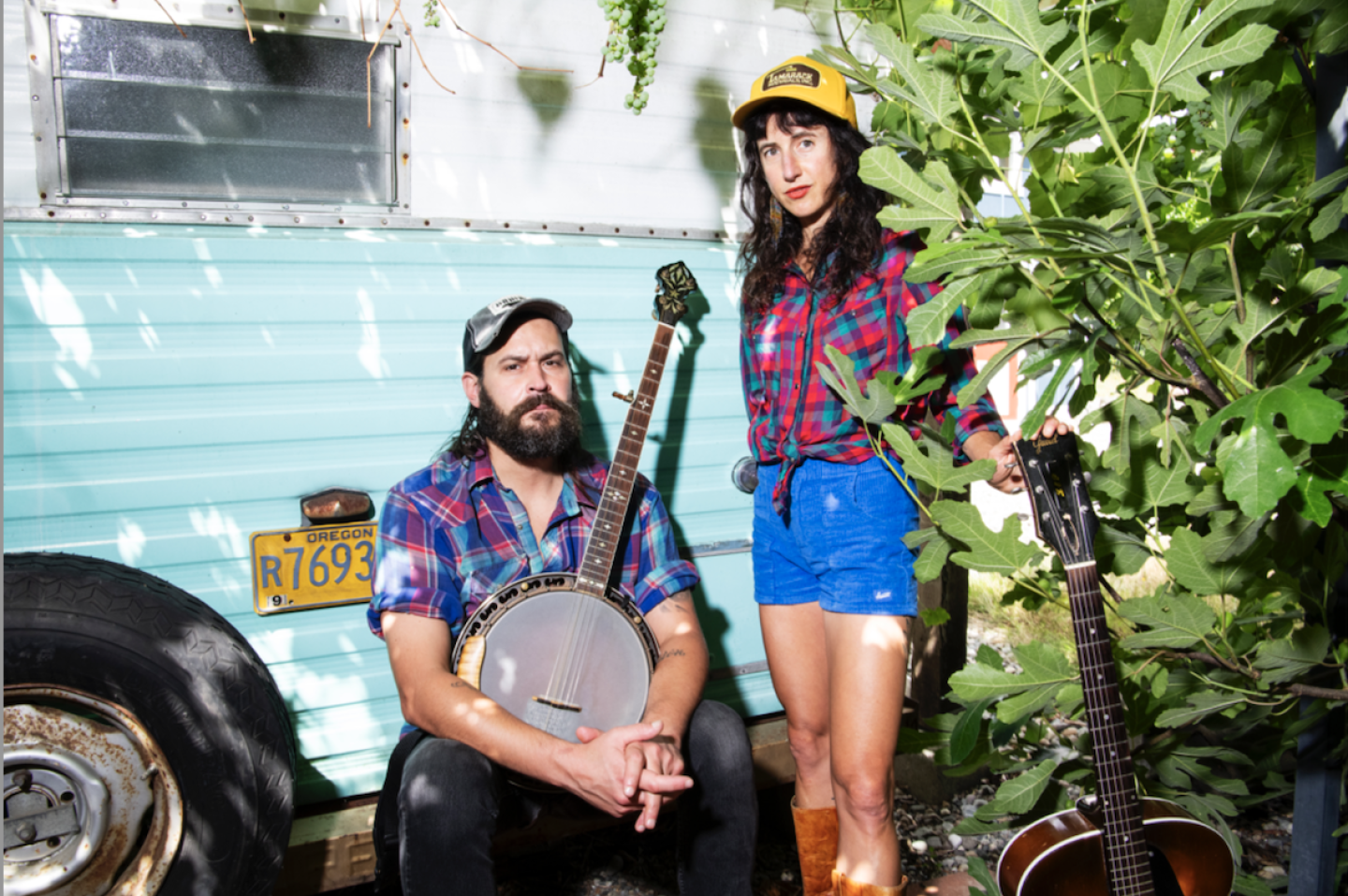 A man with a banjo and woman with a guitar pose by a vintage camper, wearing plaid shirts and trucker hats A man with a banjo and woman with a guitar pose by a vintage camper, wearing plaid shirts and trucker hats