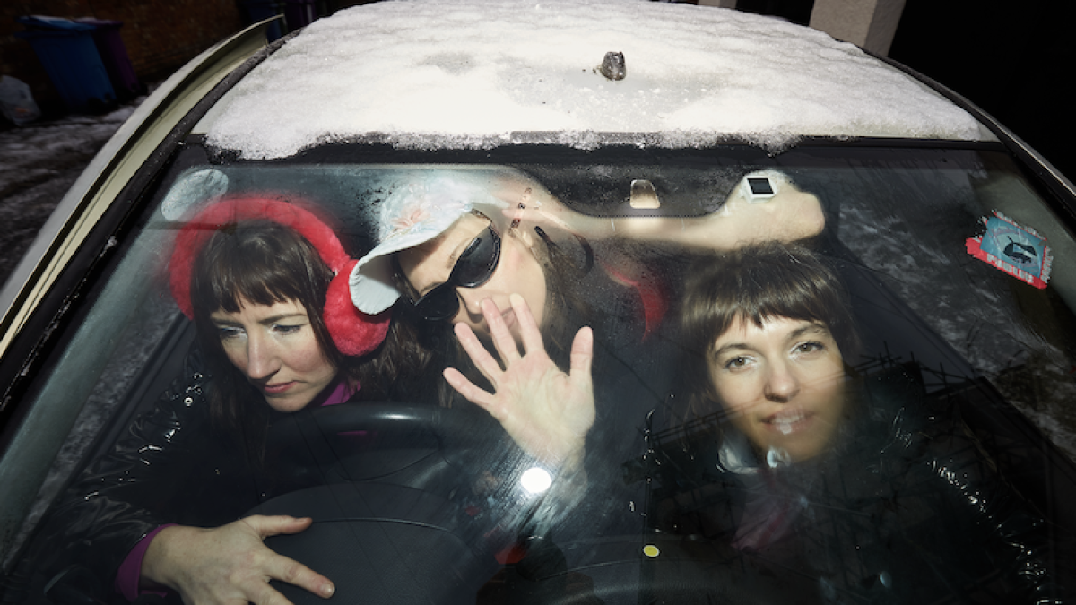 Three people are crammed inside a car with a snow-covered windshield, one driving, one waving, and one smiling at the camera.