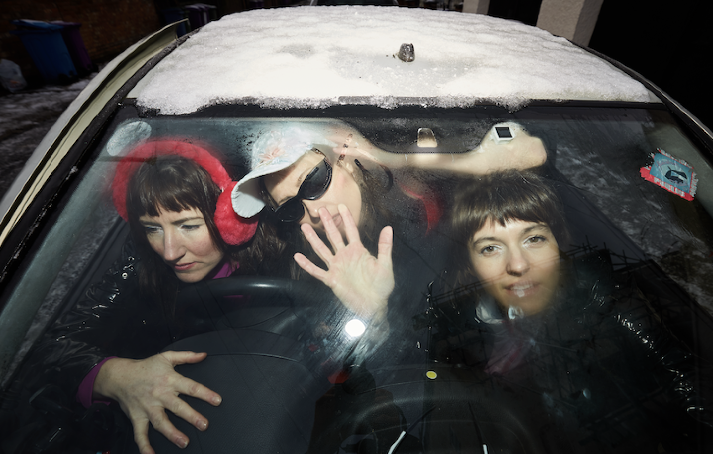 Three people are crammed inside a car with a snow-covered windshield, one driving, one waving, and one smiling at the camera. Three people are crammed inside a car with a snow-covered windshield, one driving, one waving, and one smiling at the camera.