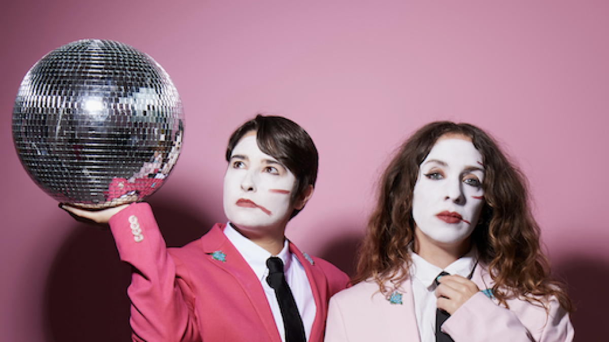 Two people in pink suits with white-painted faces pose against a pink background; one holds up a disco ball while the other adjusts their tie.