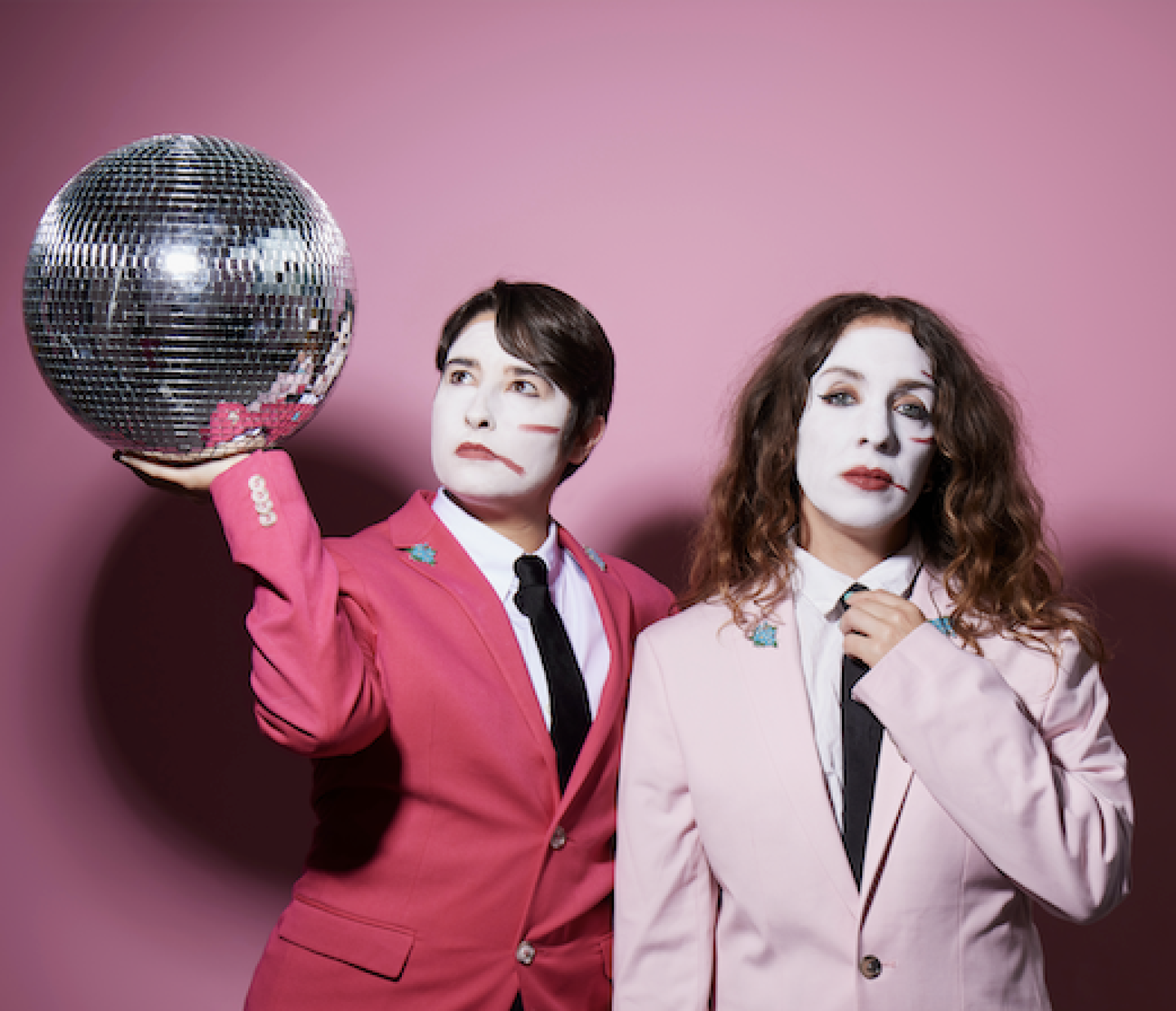 Two people in pink suits with white-painted faces pose against a pink background; one holds up a disco ball while the other adjusts their tie. Two people in pink suits with white-painted faces pose against a pink background; one holds up a disco ball while the other adjusts their tie.