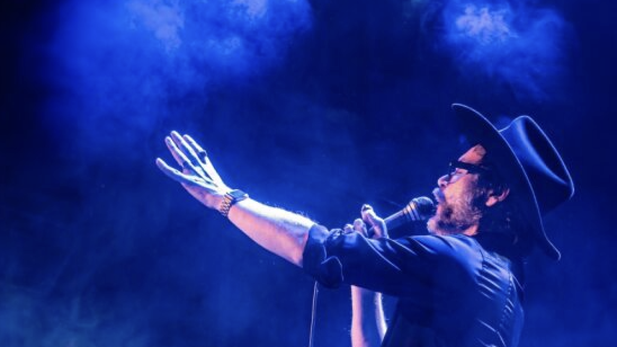 A side profile of a singer in a cowboy hat performing on stage, holding a microphone with one arm extended, surrounded by smoky blue lighting.