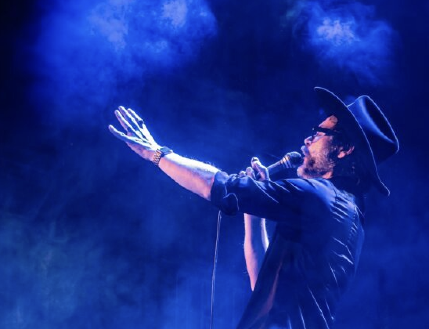 A side profile of a singer in a cowboy hat performing on stage, holding a microphone with one arm extended, surrounded by smoky blue lighting. A side profile of a singer in a cowboy hat performing on stage, holding a microphone with one arm extended, surrounded by smoky blue lighting.