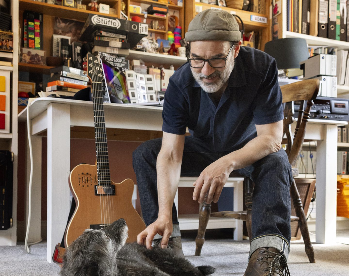 A person sat on a chair leaning down to stroke dog. Behind them shelves full of books and items. A person sat on a chair leaning down to stroke dog. Behind them shelves full of books and items.