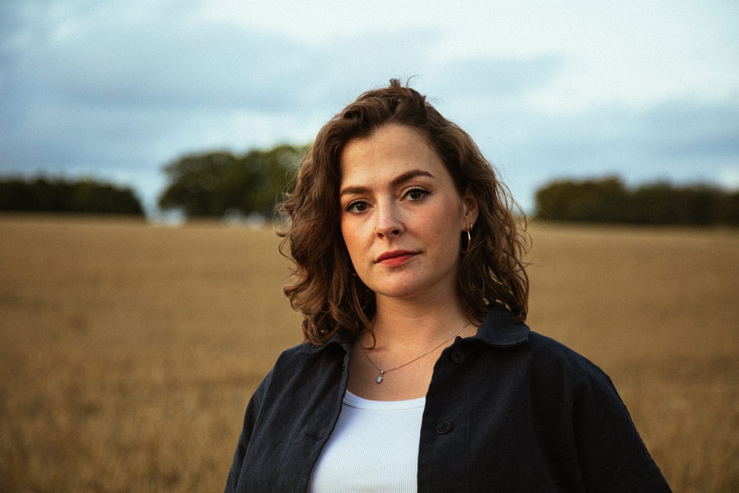 Woman with wavy brown hair wearing a dark shirt over a white top, standing in a field with trees in the background. Woman with wavy brown hair wearing a dark shirt over a white top, standing in a field with trees in the background.