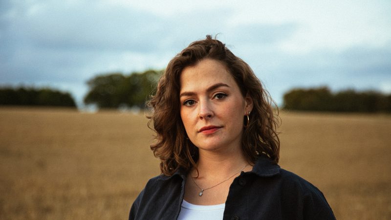 Woman with wavy brown hair wearing a dark shirt over a white top, standing in a field with trees in the background. Woman with wavy brown hair wearing a dark shirt over a white top, standing in a field with trees in the background.