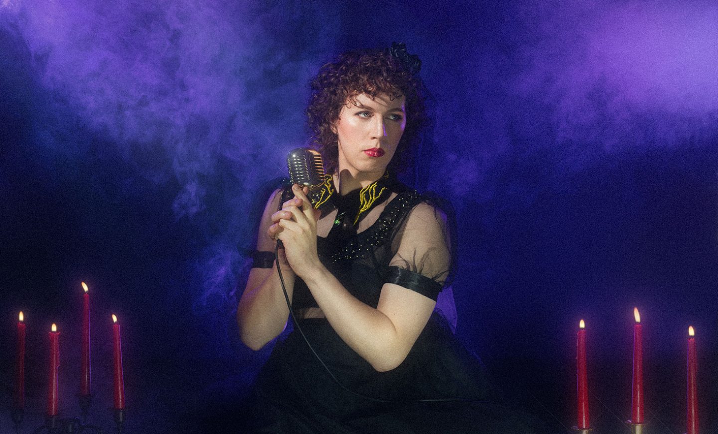 A person wearing a shear black dress and holding an old-fashioned silver microphone. They have curly brown hair and they're looking off to one side. Beside them are tall, red candles and there is a smoky purple background. A person wearing a shear black dress and holding an old-fashioned silver microphone. They have curly brown hair and they're looking off to one side. Beside them are tall, red candles and there is a smoky purple background.