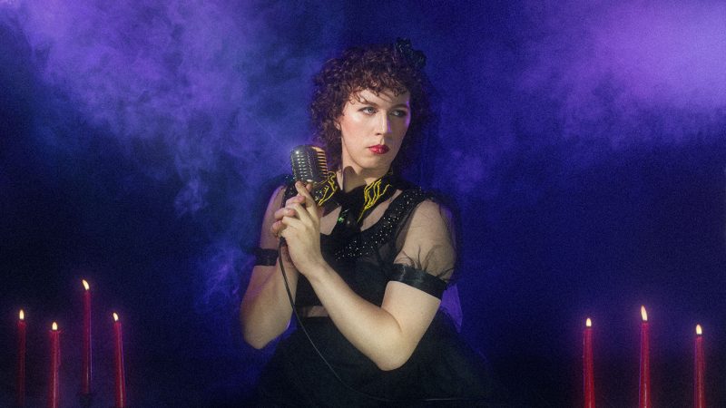 A person wearing a shear black dress and holding an old-fashioned silver microphone. They have curly brown hair and they're looking off to one side. Beside them are tall, red candles and there is a smoky purple background. A person wearing a shear black dress and holding an old-fashioned silver microphone. They have curly brown hair and they're looking off to one side. Beside them are tall, red candles and there is a smoky purple background.