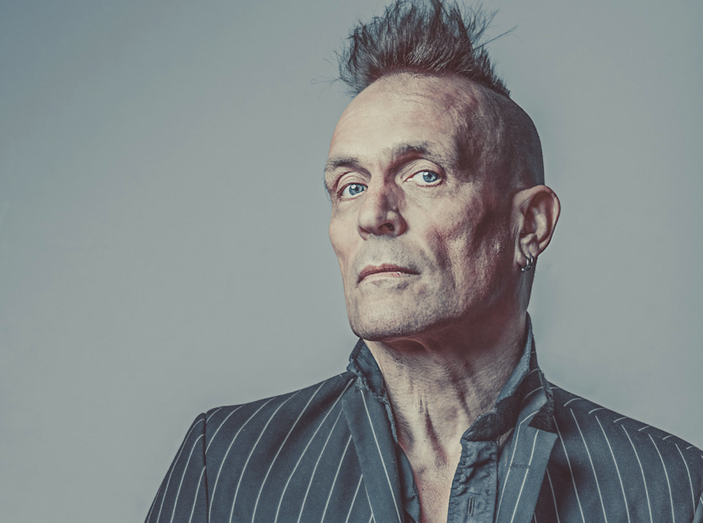 A portrait of a person with a mohawk hairstyle, light blue eyes, and a serious expression, wearing a pinstriped suit jacket and dark shirt, set against a plain grey background.